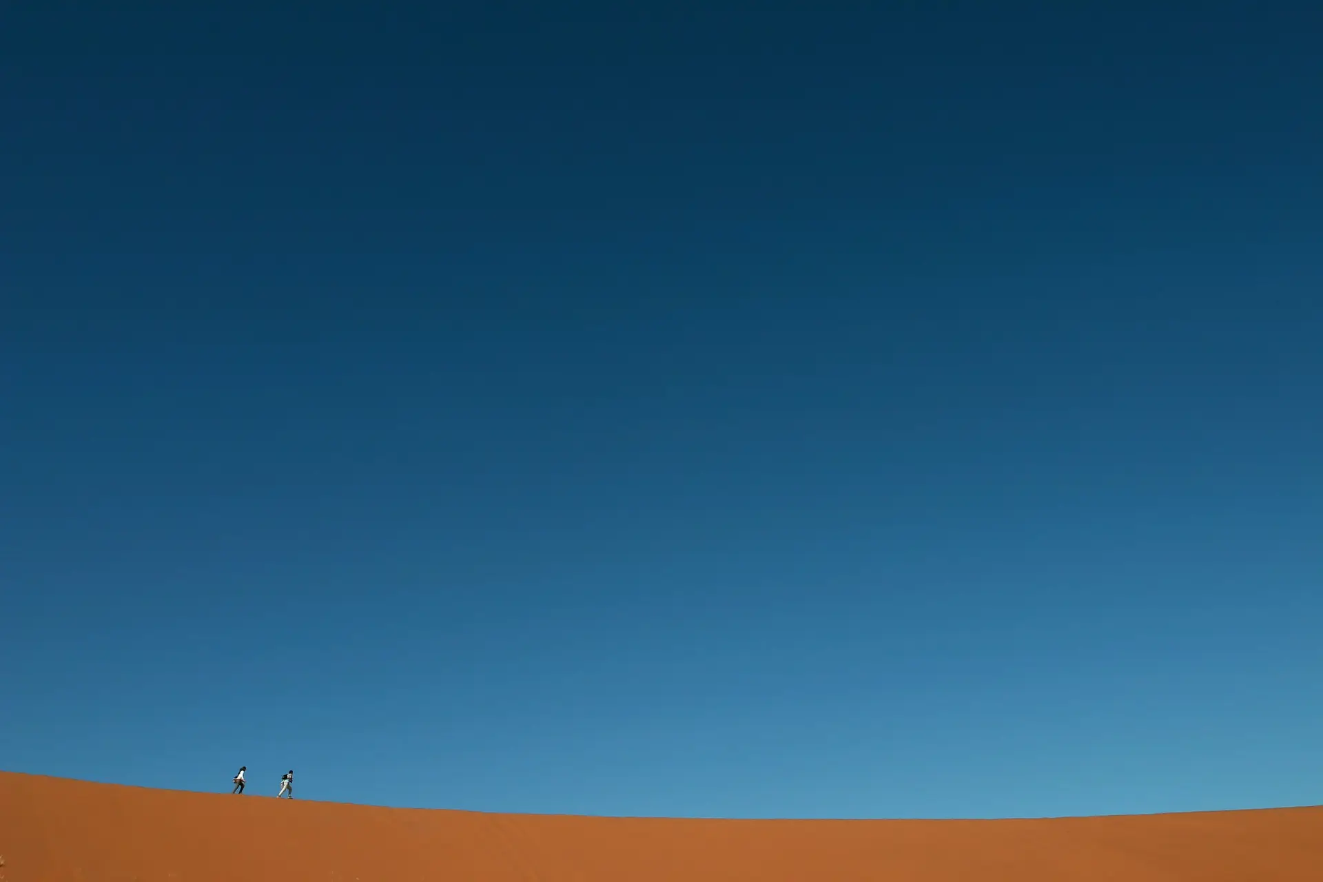 Two people walk along the crest of an orange dune under a deep blue sky